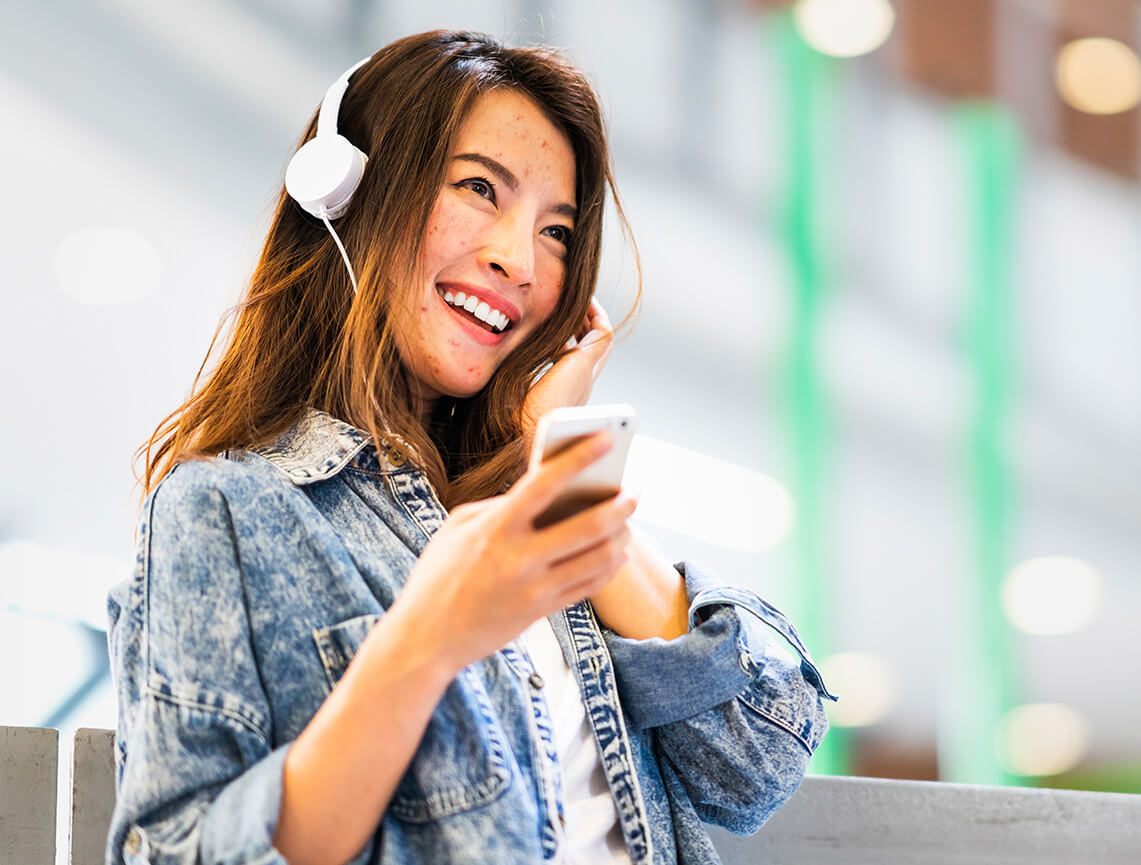 Smiling teen girl with headphones holding cell represents finding answers to FAQs on how AKLIEF® Cream works to treat acne